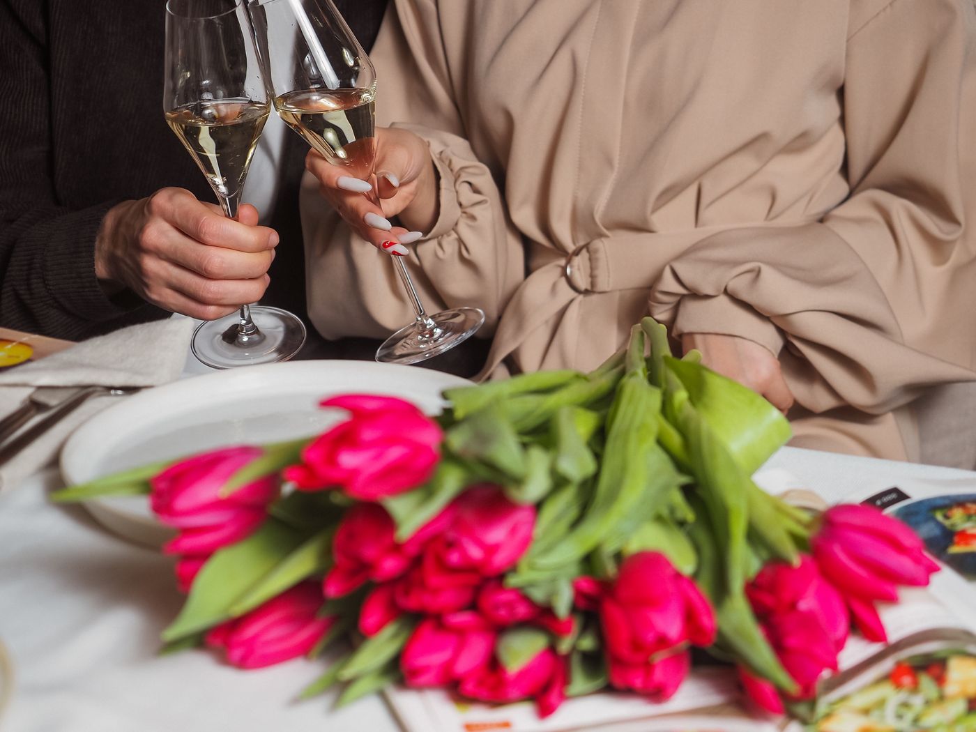 A dinner table set for two, hands raising wine glasses beside a bunch of pink tulips